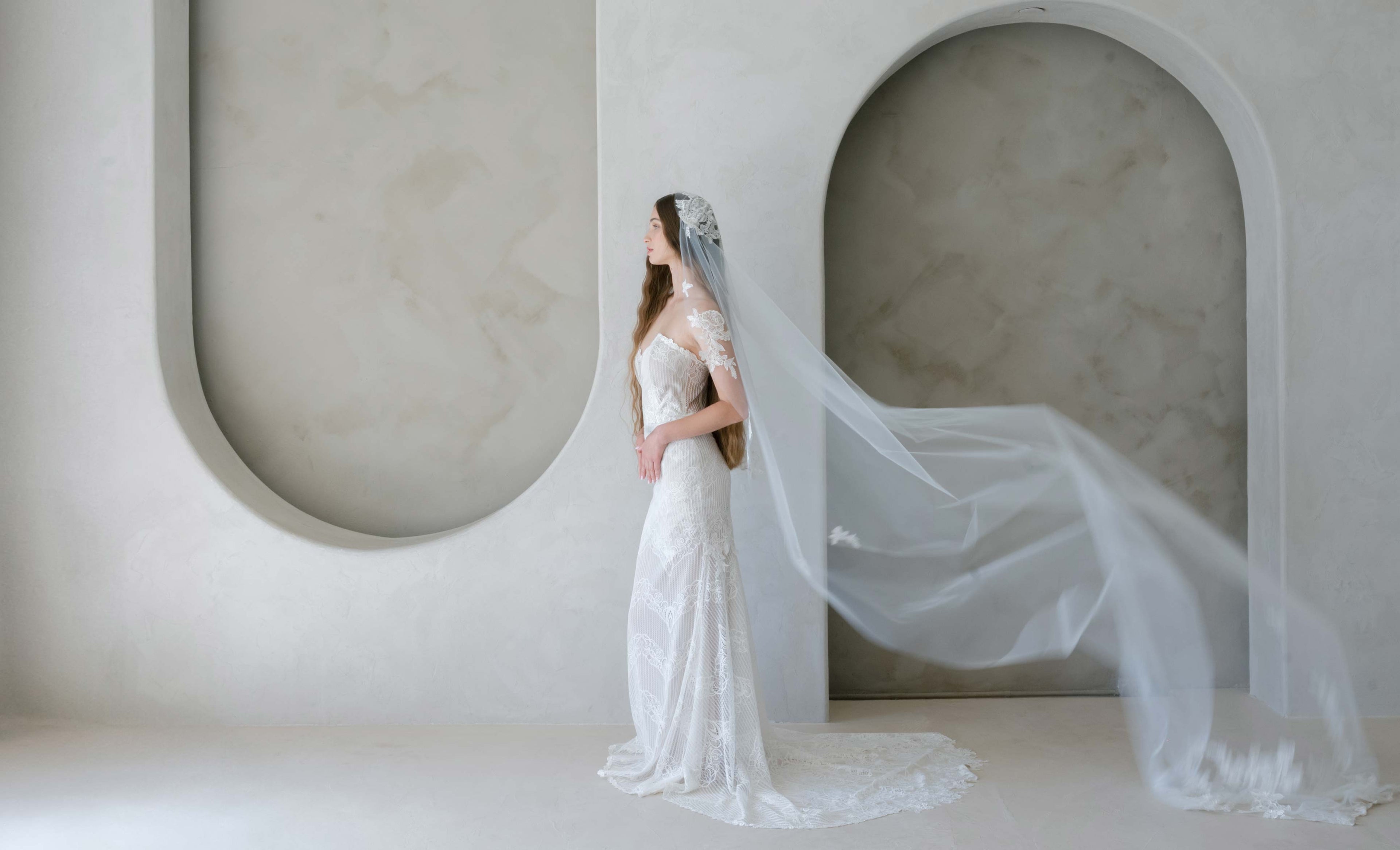 Woman in a white wedding dress with a long veil standing against a modern architectural background.