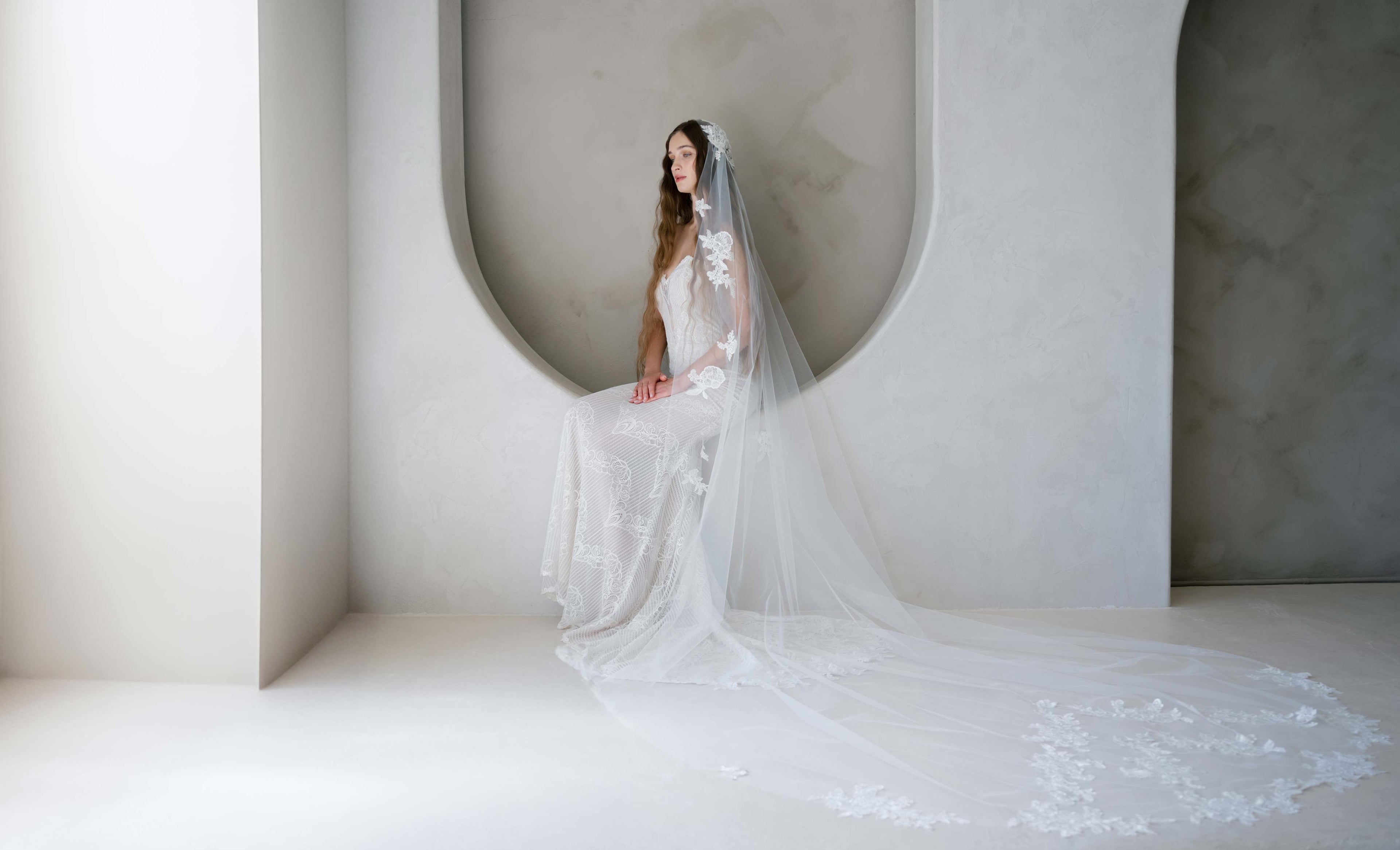 Woman in a white wedding dress with a long veil standing in a minimalist setting.