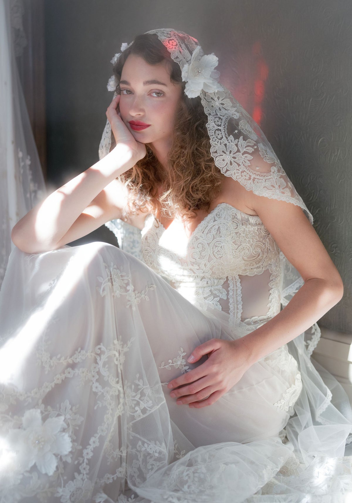 Bride seated by a window wearing the Sonnet lace mantilla veil with silk flower accents