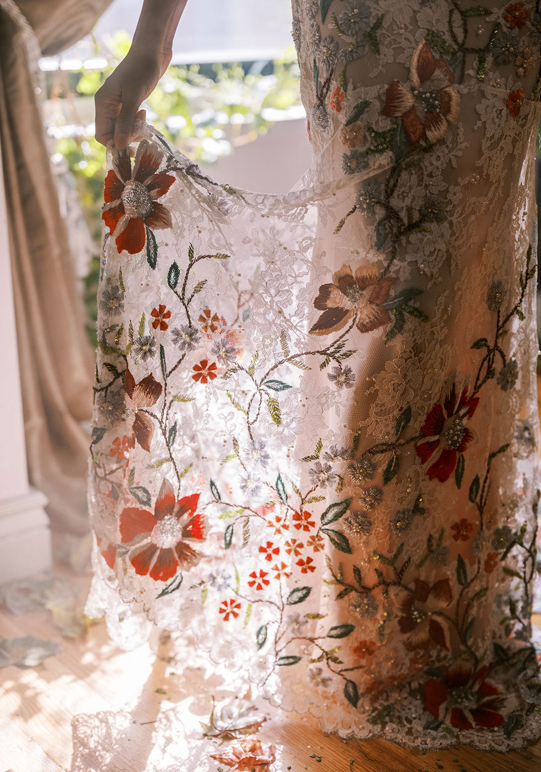 Bride holds the hem of her exquisite gown with rich red and pastel floral embroidery, shining in sunlight