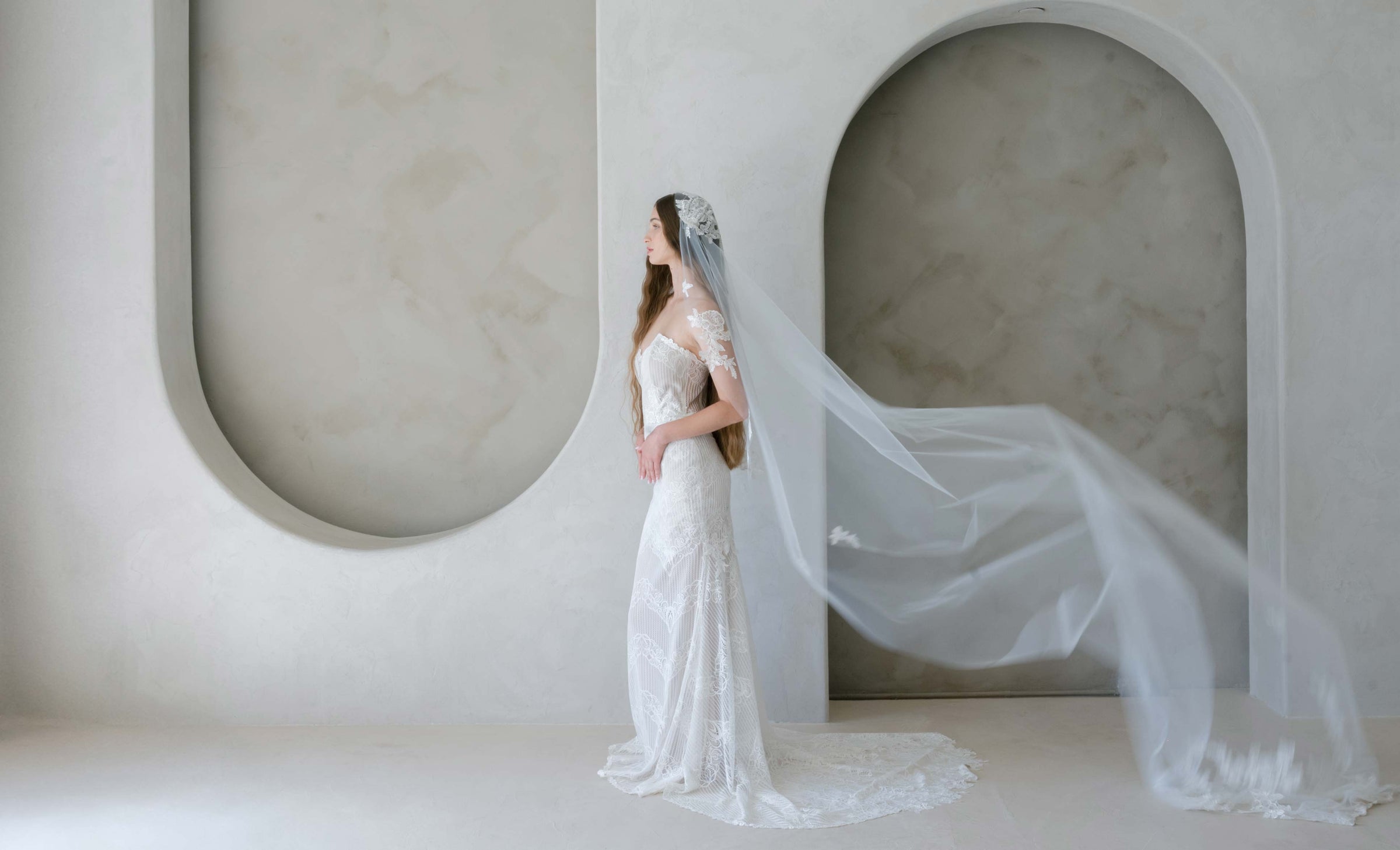Woman in a white wedding dress with a long veil standing against a modern architectural background.