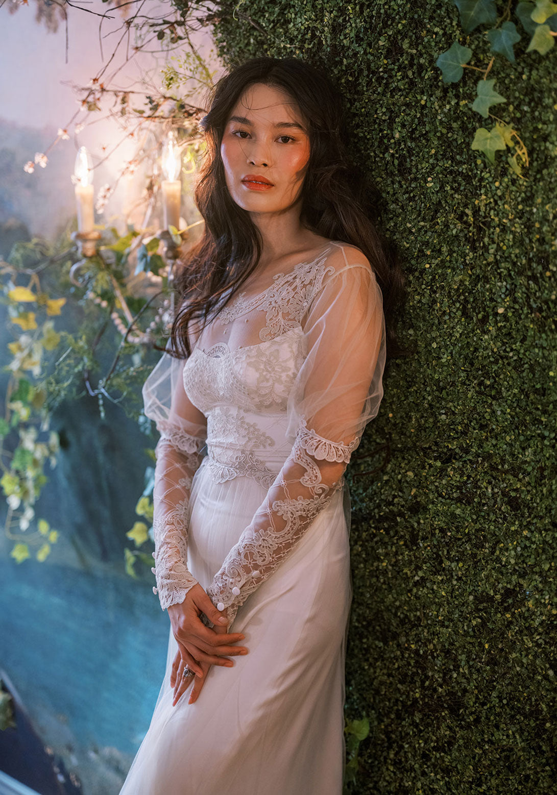 Bride leaning on green ivy wall in a romantic Trellis lace gown with elegant embroidery and sheer details.