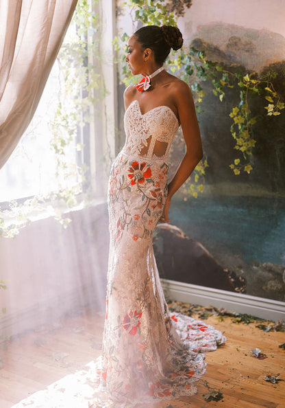 Bride in an embroidered lace gown with rich red and green florals, bathed in golden sunlight by the window.