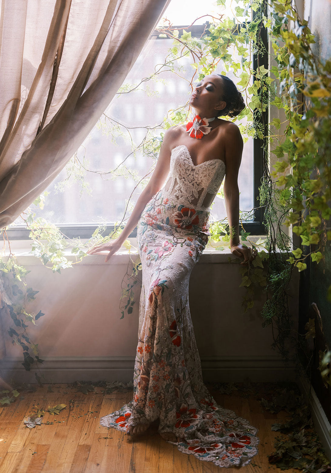 Bride sits on a windowsill in a luxe lace gown, adorned with red and pastel floral embroidery, glowing in soft light